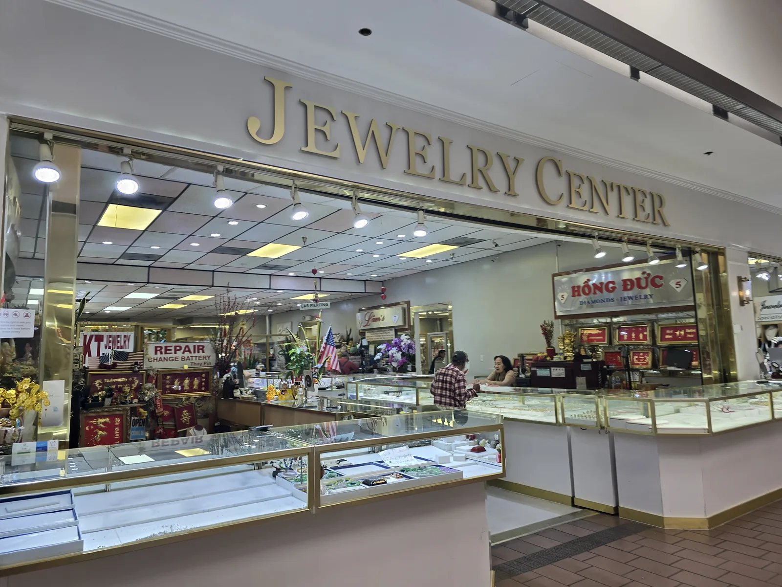 Jewelry Center 内景,多个独立柜台如 Hong Duc、KT Jewelry,墙上有 Repair Change Battery 与美国国旗